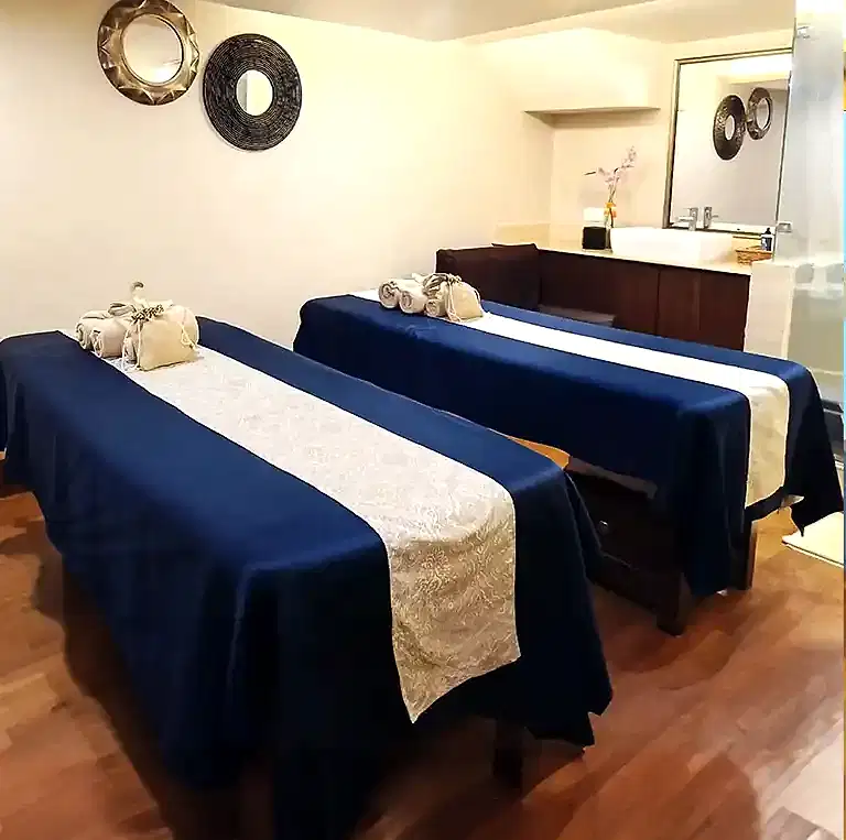 Elegant couple massage room at Tattva Wellness Spa Novotel Imagica Khopoli with two massage beds draped in navy-blue and beige linens, warm wooden flooring, soft wall lighting, and soothing décor that promotes deep relaxation.