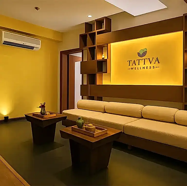Warmly lit lounge at Tattva Wellness Spa, Royal Orchid Resort Goa, showcasing a cozy beige sofa, wooden decor accents, and a soft yellow backdrop creating a serene pre-therapy ambiance.