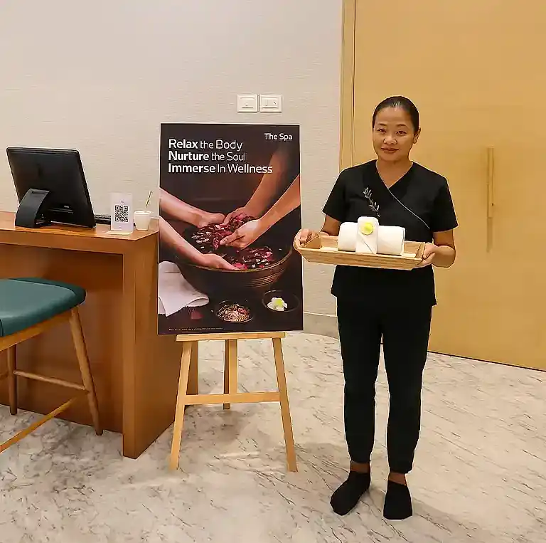 Reception area at Tattva Spa, Fairfield by Marriott, featuring a spa professional holding a welcome tray beside a wellness-themed poster promoting relaxation and rejuvenation.