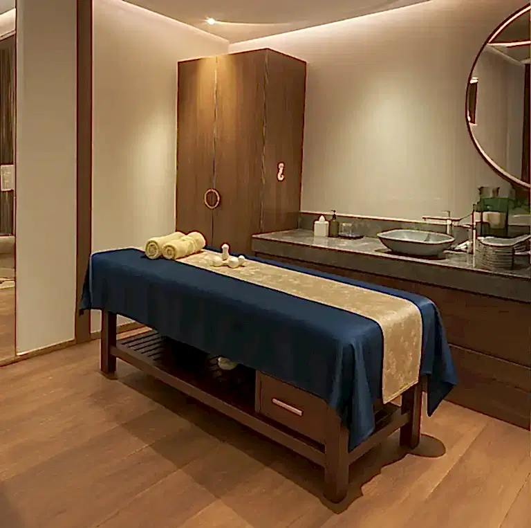 Warm-lit massage room at Tattva Wellness Spa, Novotel Mumbai International Airport, featuring a neatly arranged therapy bed with blue and gold linen and wooden interiors.