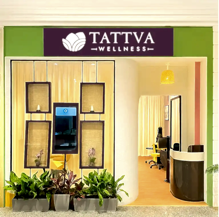 Entrance of Tattva Wellness Spa at Manohar International Airport Goa, featuring a bright green and lavender facade with indoor plants and a welcoming reception visible through the glass doorway.